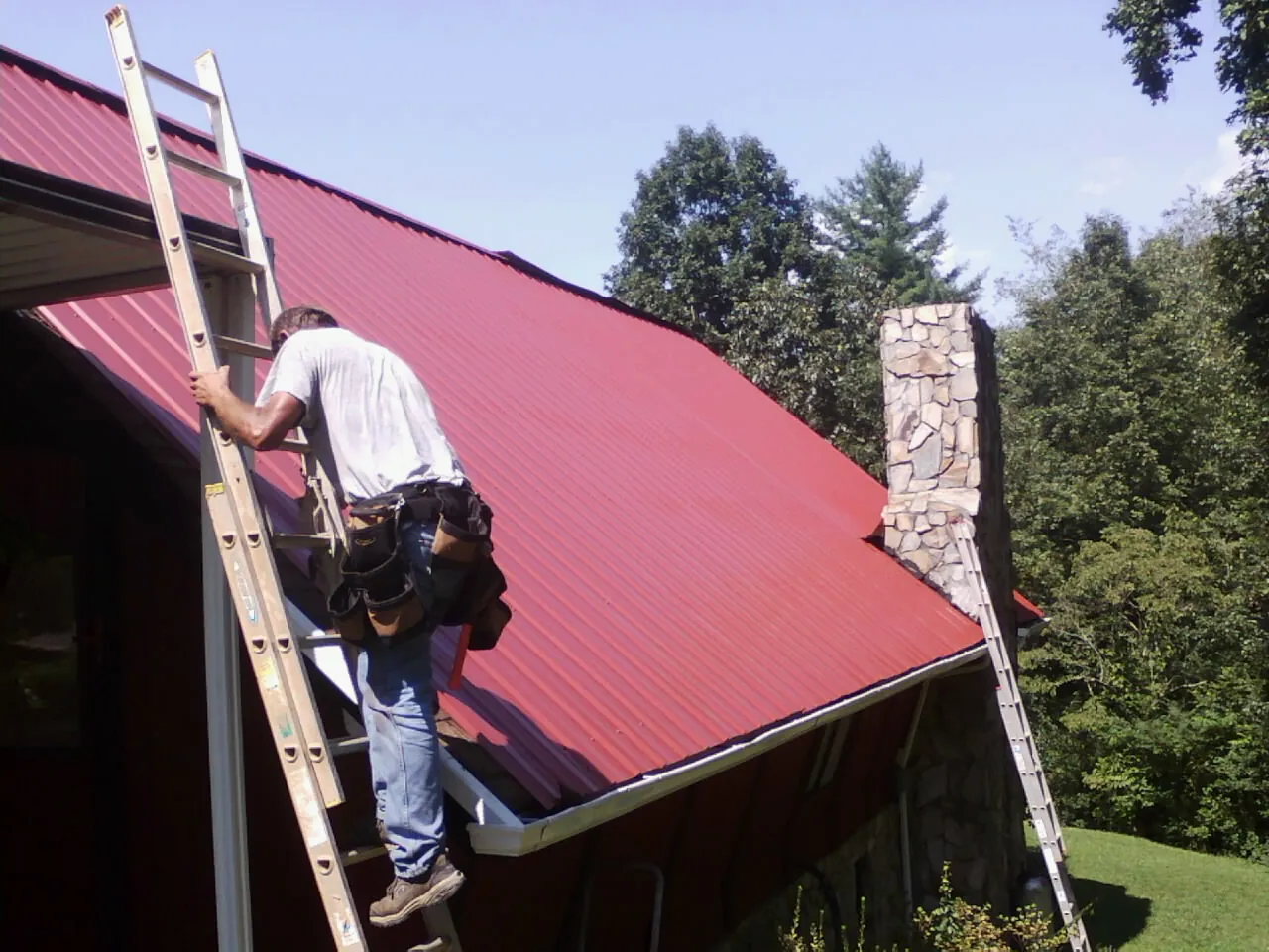 Roofer on a ladder beside a red metal roof and stone chimney — Soffit & Fascia Repair by Roofing Brothers in Willingboro